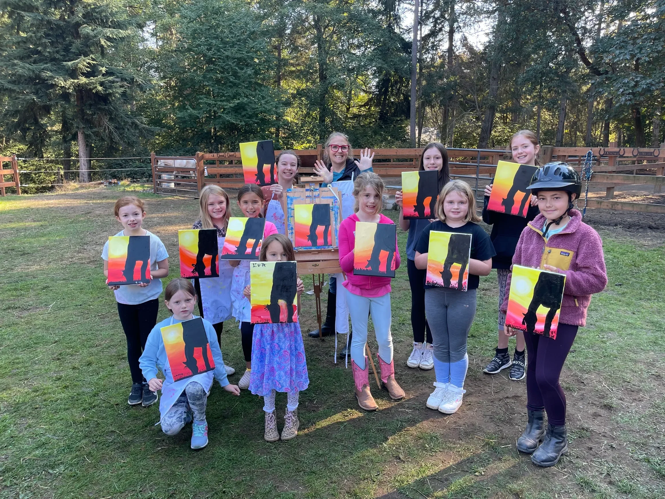 Group of children and a woman outdoors in a park holding paintings of a sunset and silhouette of an elephant.