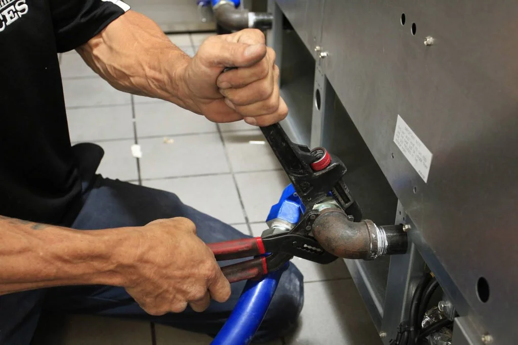 A person wearing a black shirt and blue pants is using pliers to tighten a pipe connection on a large metal appliance, possibly an HVAC or industrial equipment, in a tiled room.