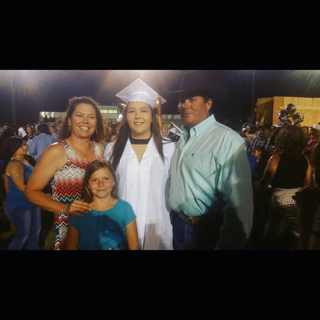 A family celebrating a graduation outdoors at night, with a young woman in a graduation cap and gown, surrounded by adults and a girl, all smiling.
