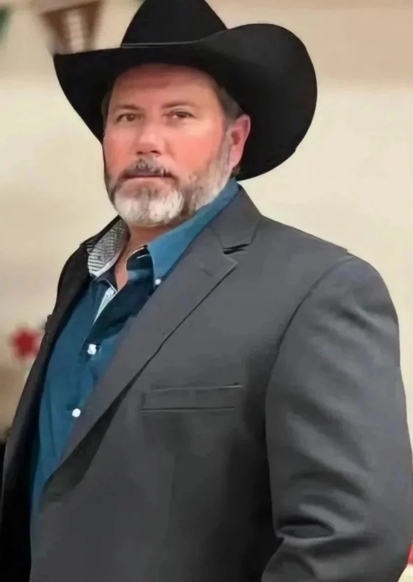 A man with a beard wearing a black cowboy hat, a dark gray suit jacket, and a blue collared shirt, standing indoors