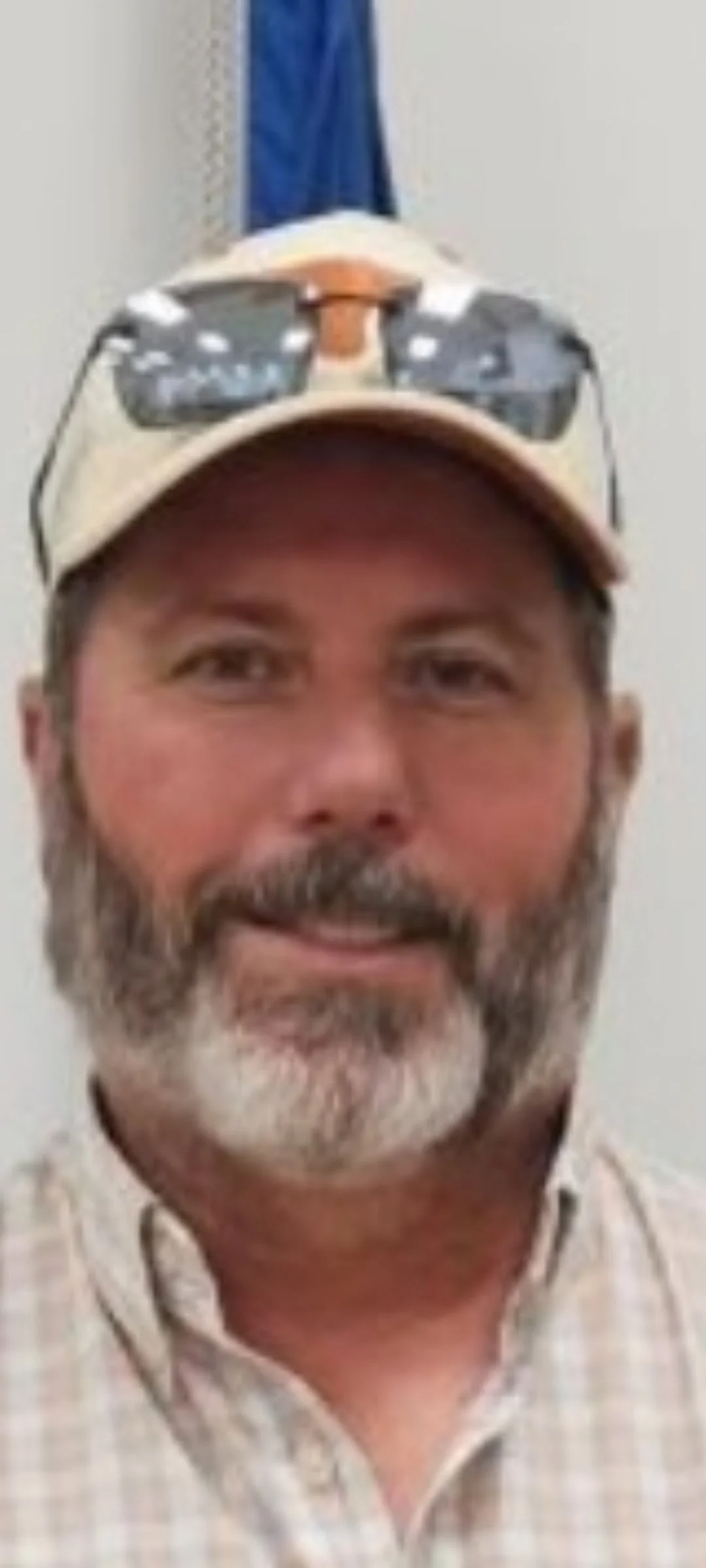A man with a beard wearing a beige baseball cap with sunglasses on top, a checkered shirt, standing in front of a light-colored background.