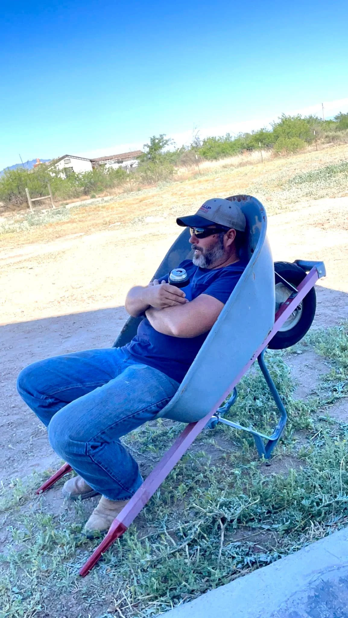 A man sitting in a wheelbarrow outdoors, wearing sunglasses, a baseball cap, a navy blue shirt, jeans, and brown boots, holding a can. The background shows a dirt road, some bushes, and houses in the distance under a clear blue sky.