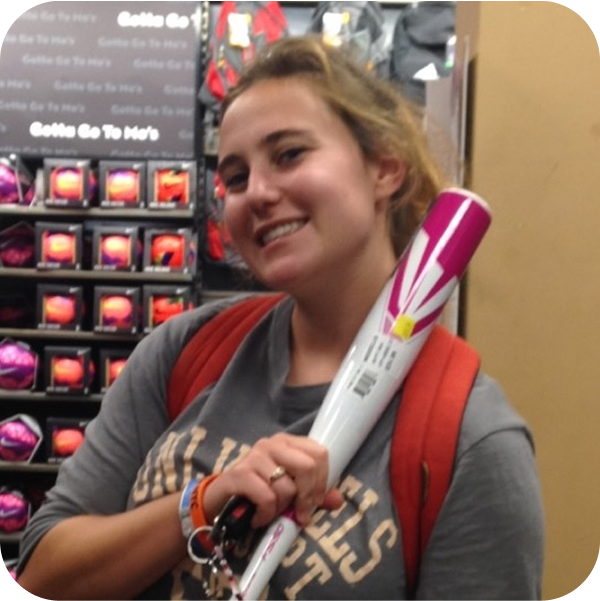 A young woman with a smile, holding a rolled-up poster or paper in a store aisle. She wears a gray T-shirt with text, a backpack, and has a colorful wristband.