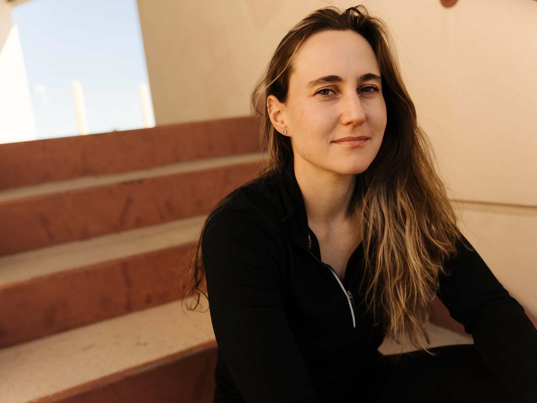 A young woman with long, wavy brown hair, light skin, and wearing a black zip-up top sits on wooden stairs with a neutral expression.