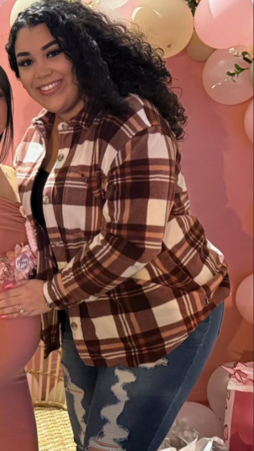 Young woman with curly black hair smiling, wearing a brown and beige plaid shirt and ripped jeans, at a celebration with pink balloons and decorations.