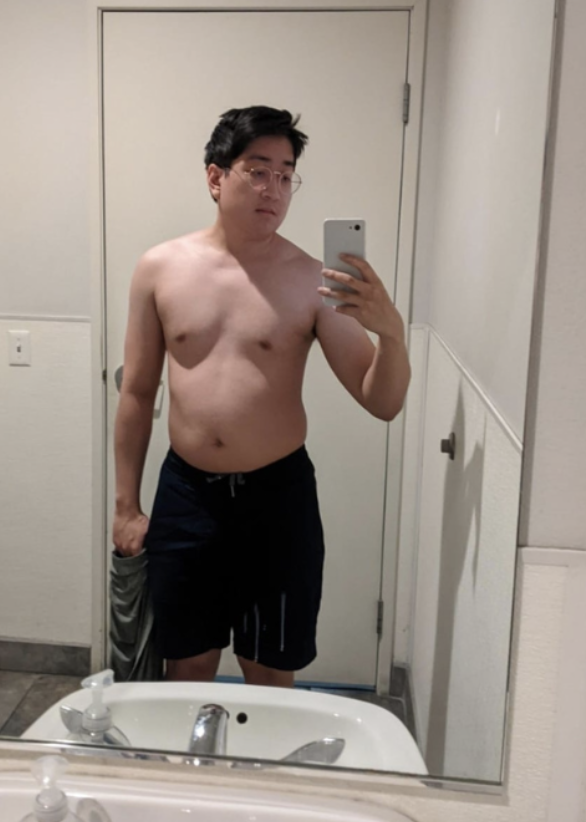 A man taking a mirror selfie in a bathroom, shirtless, wearing black shorts, holding a gray shirt in his left hand, with glasses and dark hair.