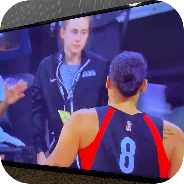 A basketball player wearing a black and red jersey with the number 8 is talking to a woman in a dark jacket in front of a television screen at a sports event.