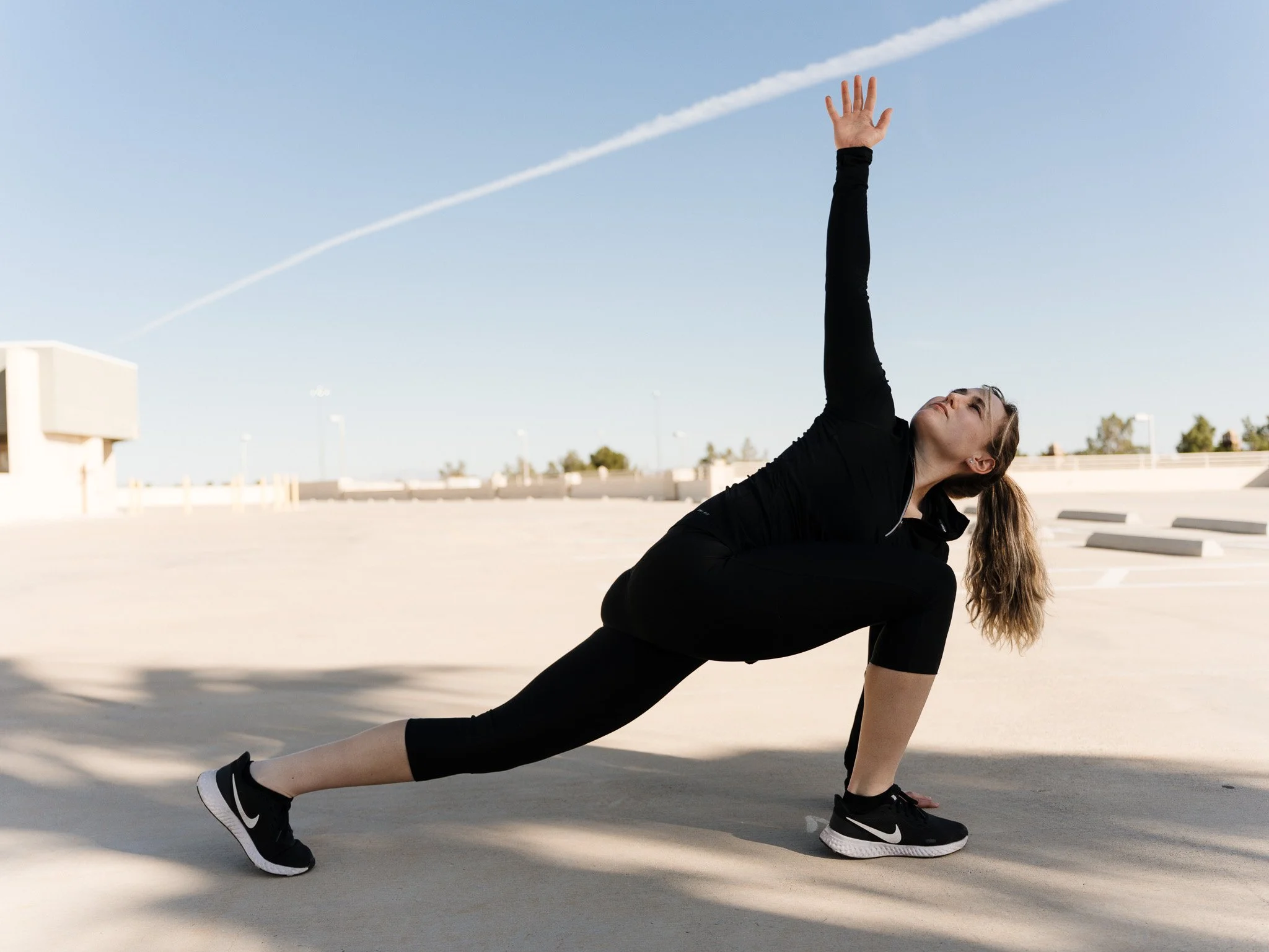 A woman in black athletic clothing and sneakers performing a yoga pose in an outdoor parking lot on a sunny day.