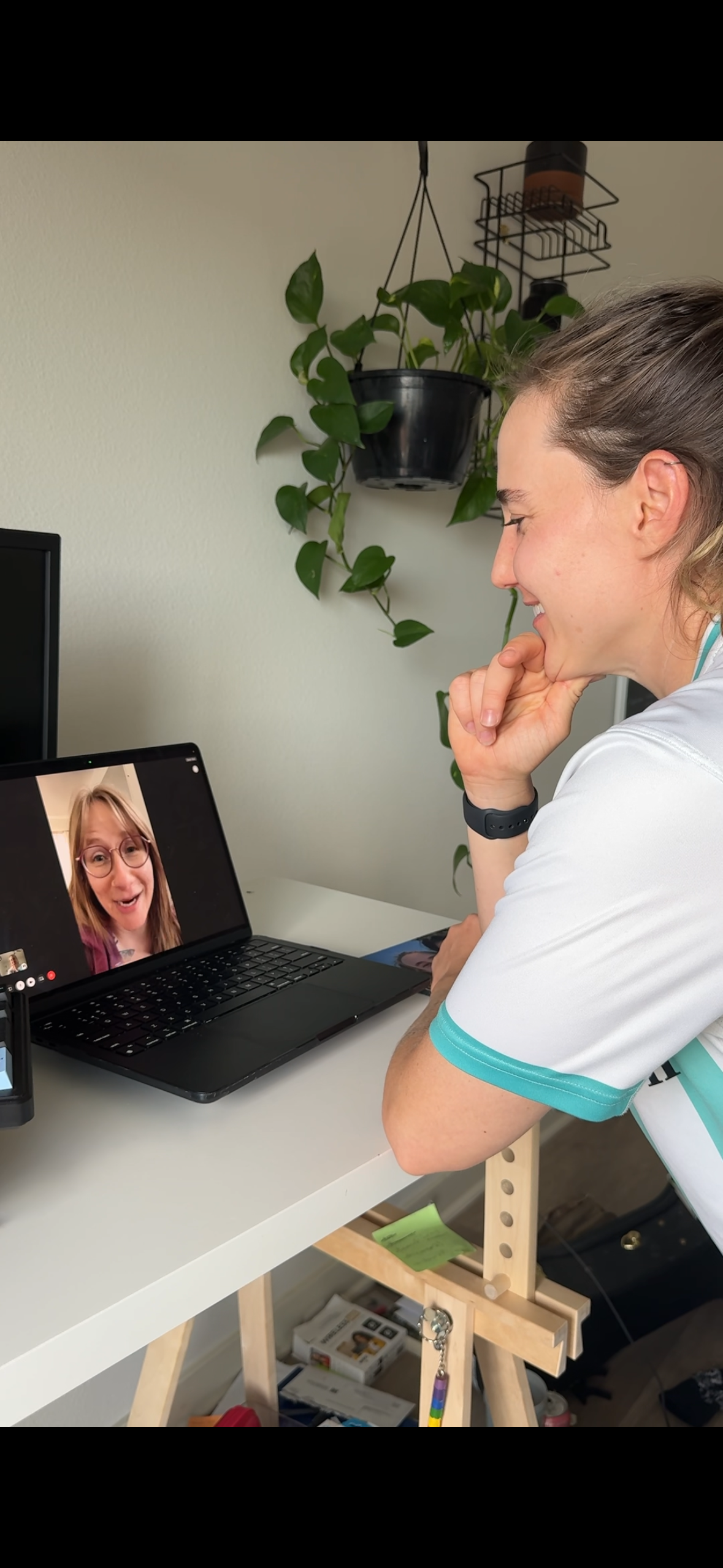 A woman with a ponytail smiling and talking on a video call with a woman on a laptop. The woman on the call is wearing glasses and a purple top. The setting is a home office with a plant and shelf in the background.
