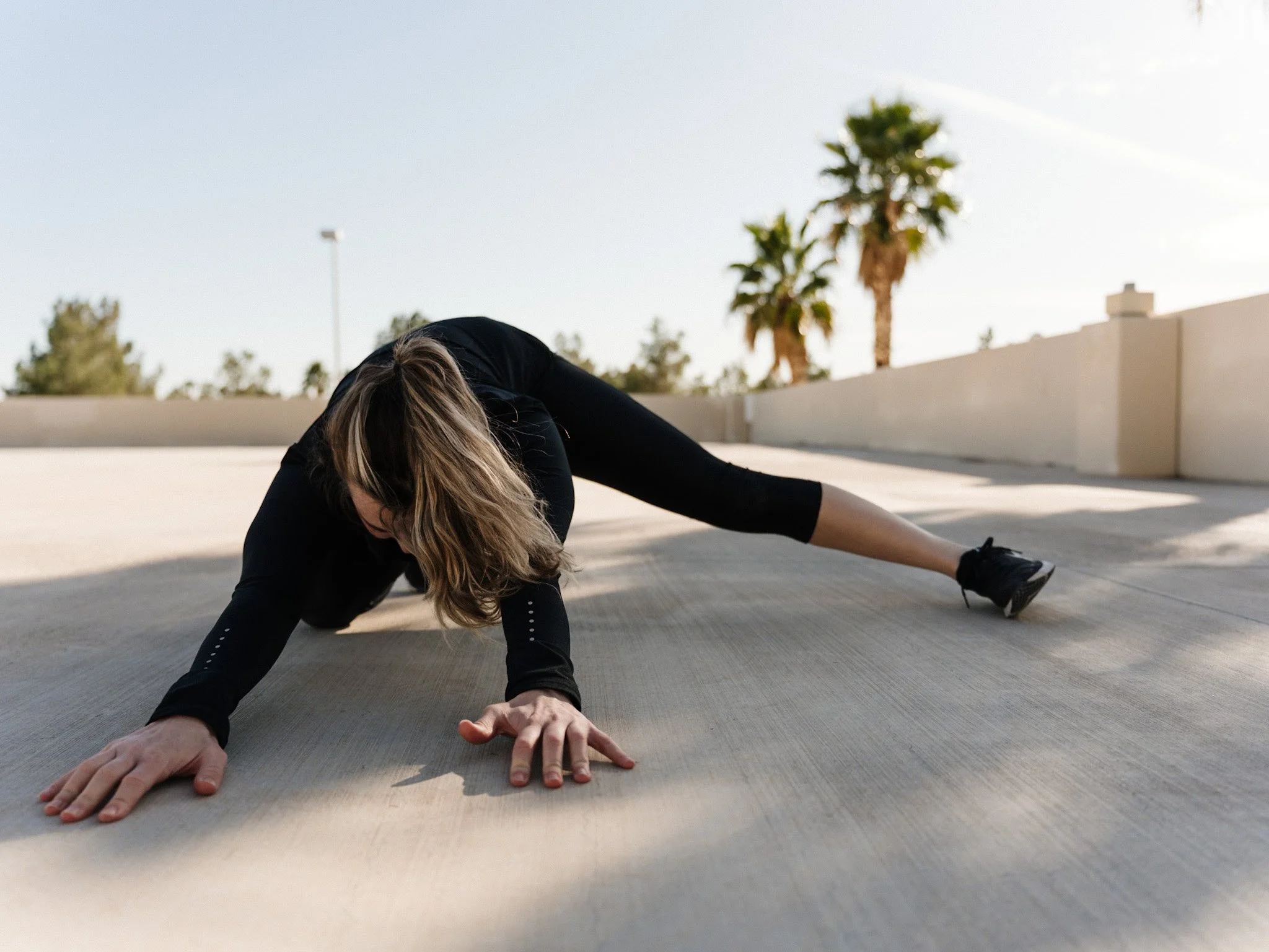 Woman in black workout clothes doing a stretching exercise on an outdoor concrete surface with palm trees and a beige wall in the background.