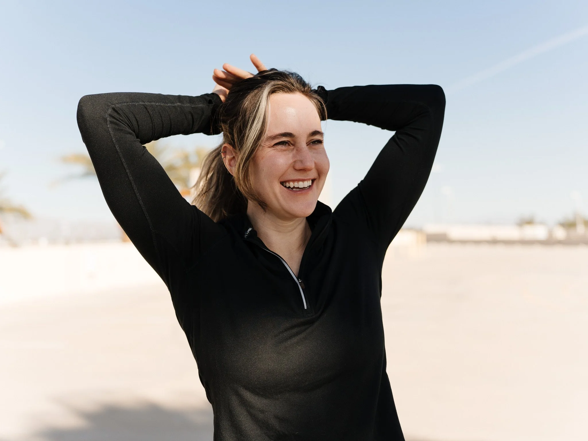 A smiling woman with long hair, wearing a black athletic top, standing on a beach with palm trees, sand, and a blue sky in the background.
