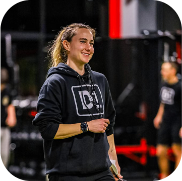 A young woman coaching and wearing a black hoodie inside a gym, with other people working out in the background.