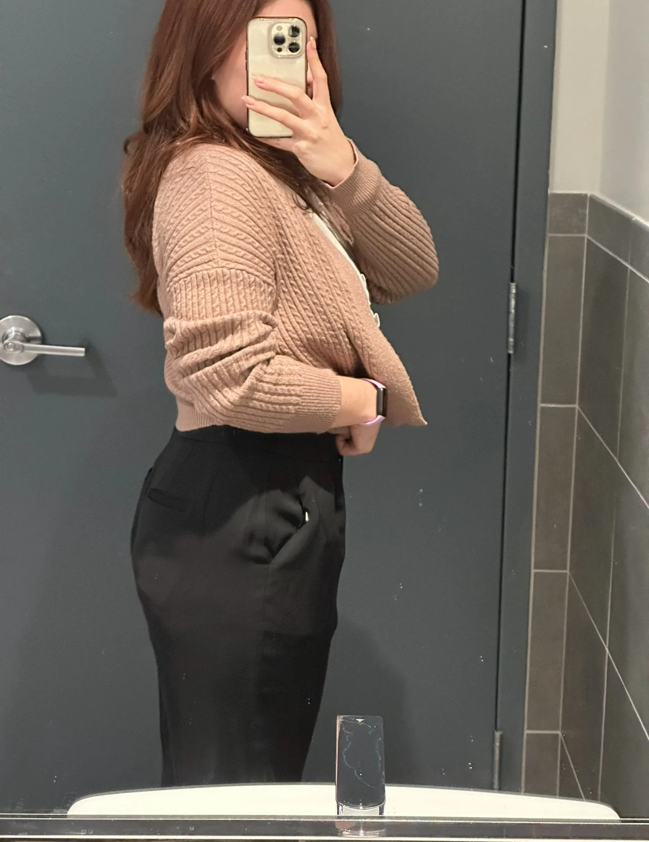 A woman taking a mirror selfie in a bathroom, wearing a tan knit sweater and black pants, with her phone covering part of her face.