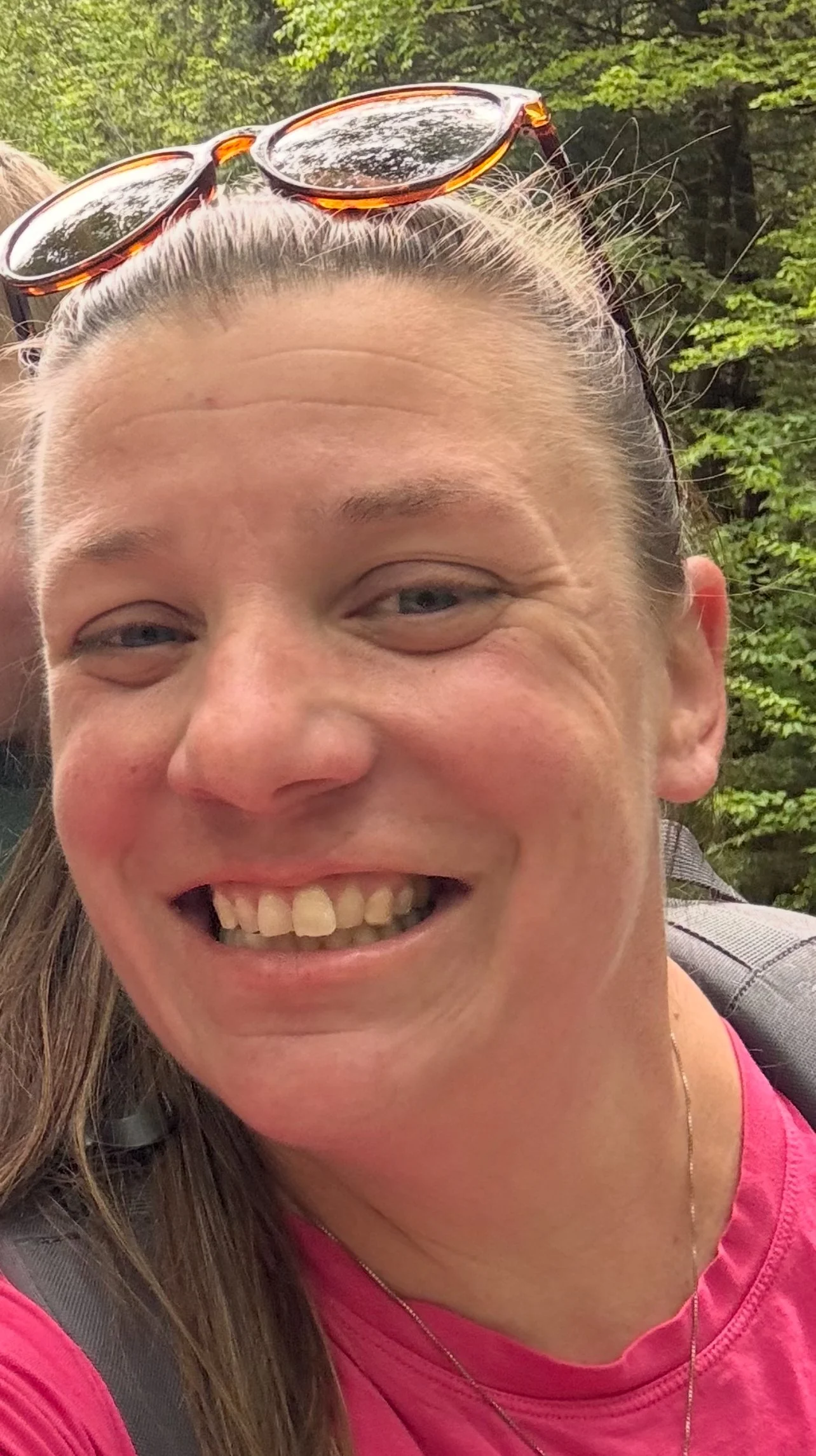 Close-up of a woman with sunglasses on her head, smiling, in a natural outdoor setting with green trees in the background.