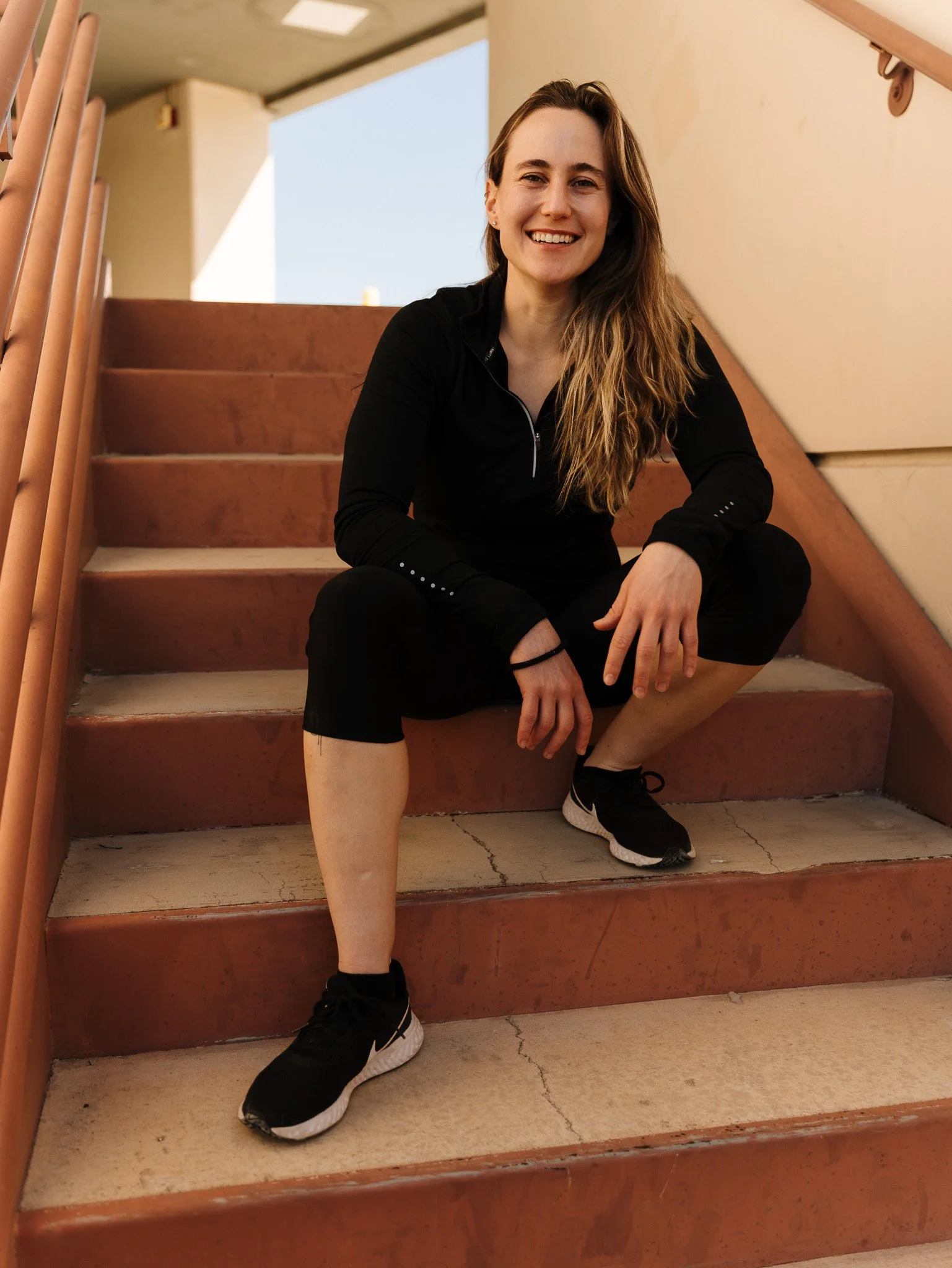 Young woman sitting on outdoor staircase, smiling, wearing black athletic wear and sneakers.