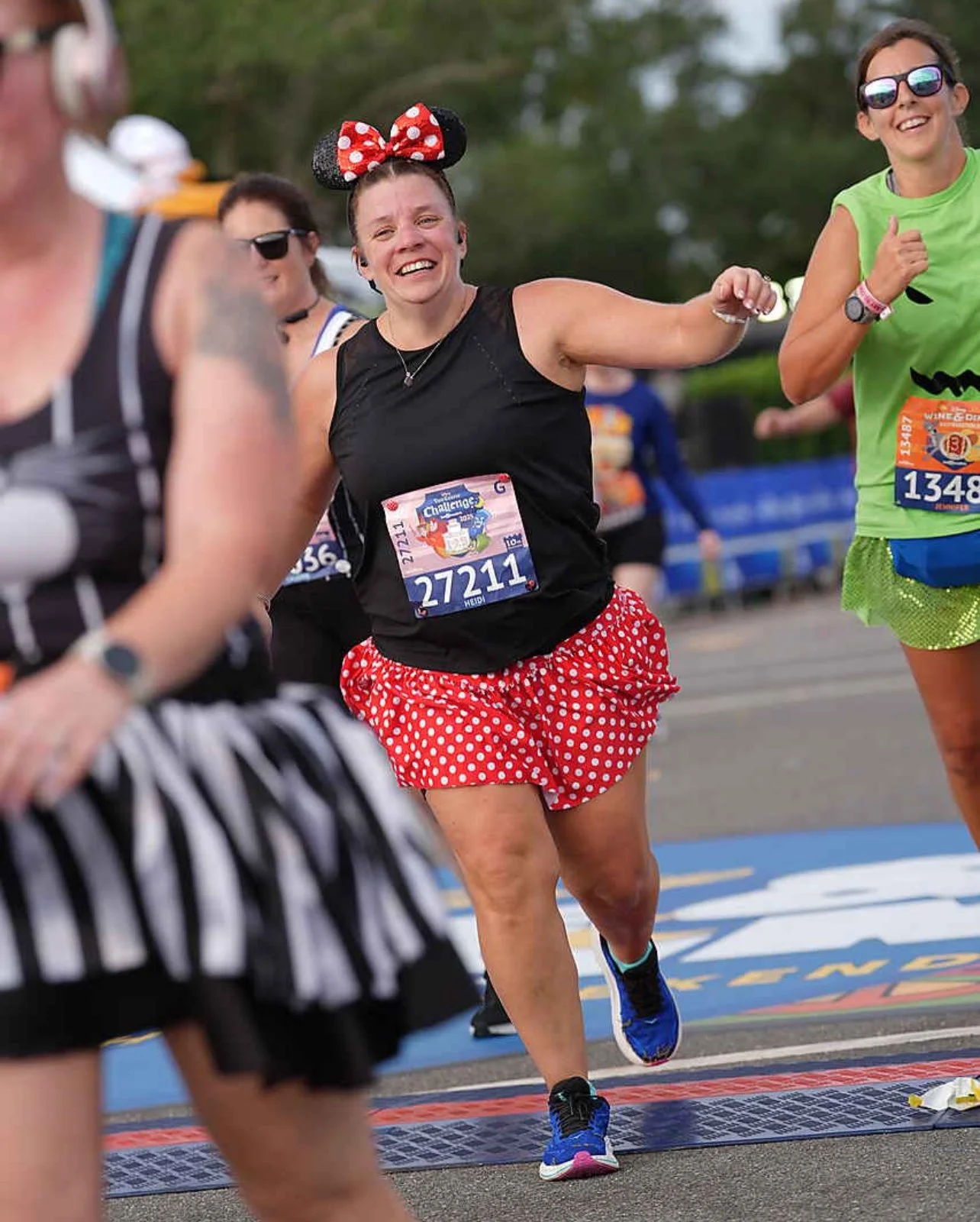 A group of runners participating in a race, with one woman in a Minnie Mouse-themed outfit, wearing a bow headband, black top, and red polka dot shorts, smiling as she runs.