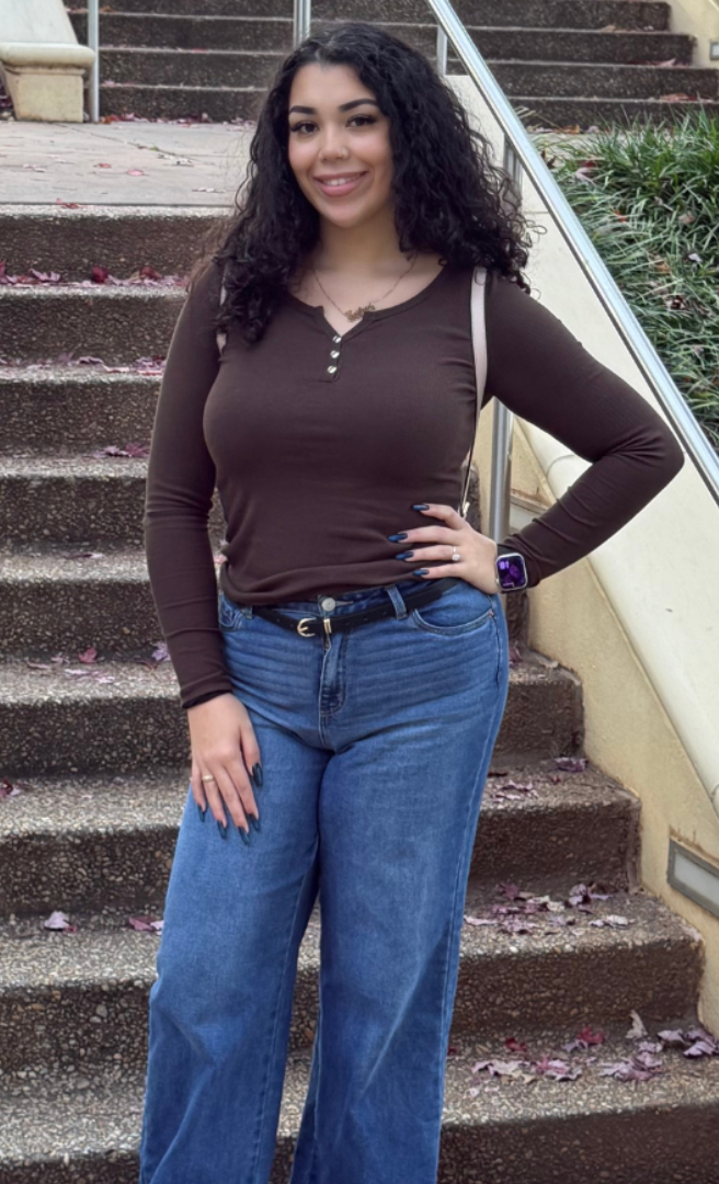 A young woman with curly black hair smiling, standing outside on a set of stairs, wearing a long-sleeve brown top, blue jeans, and a smartwatch, with pink petals on the ground.