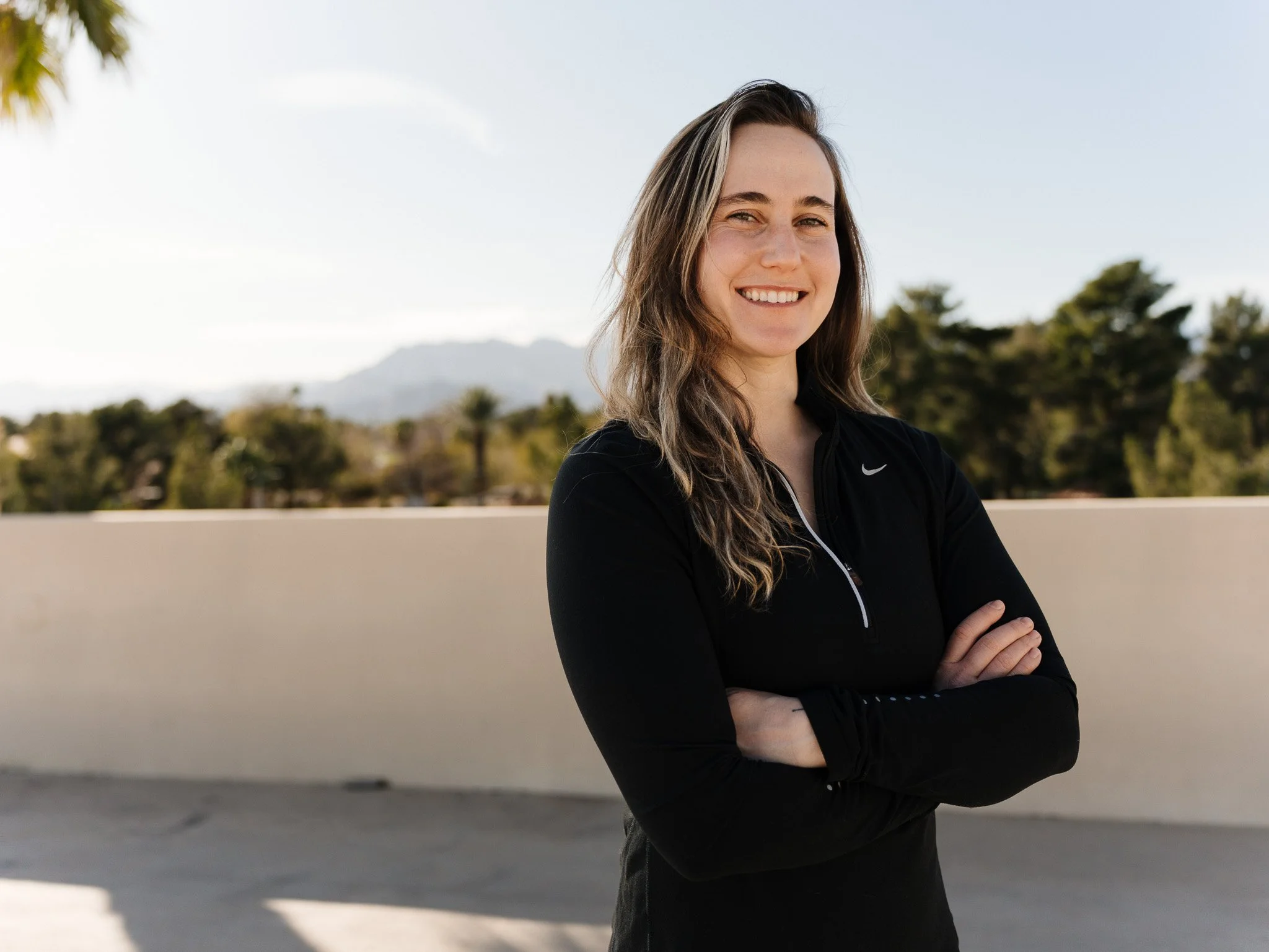 A woman with long wavy hair smiling, wearing a black Nike athletic top with a quarter zipper, standing outdoors with trees and mountains in the background.