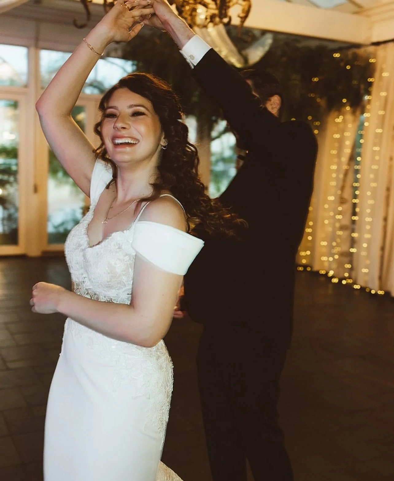 A bride and groom dancing at their wedding reception, with the bride smiling and the groom's face partially obscured, surrounded by fairy lights and a bright, elegant indoor venue.