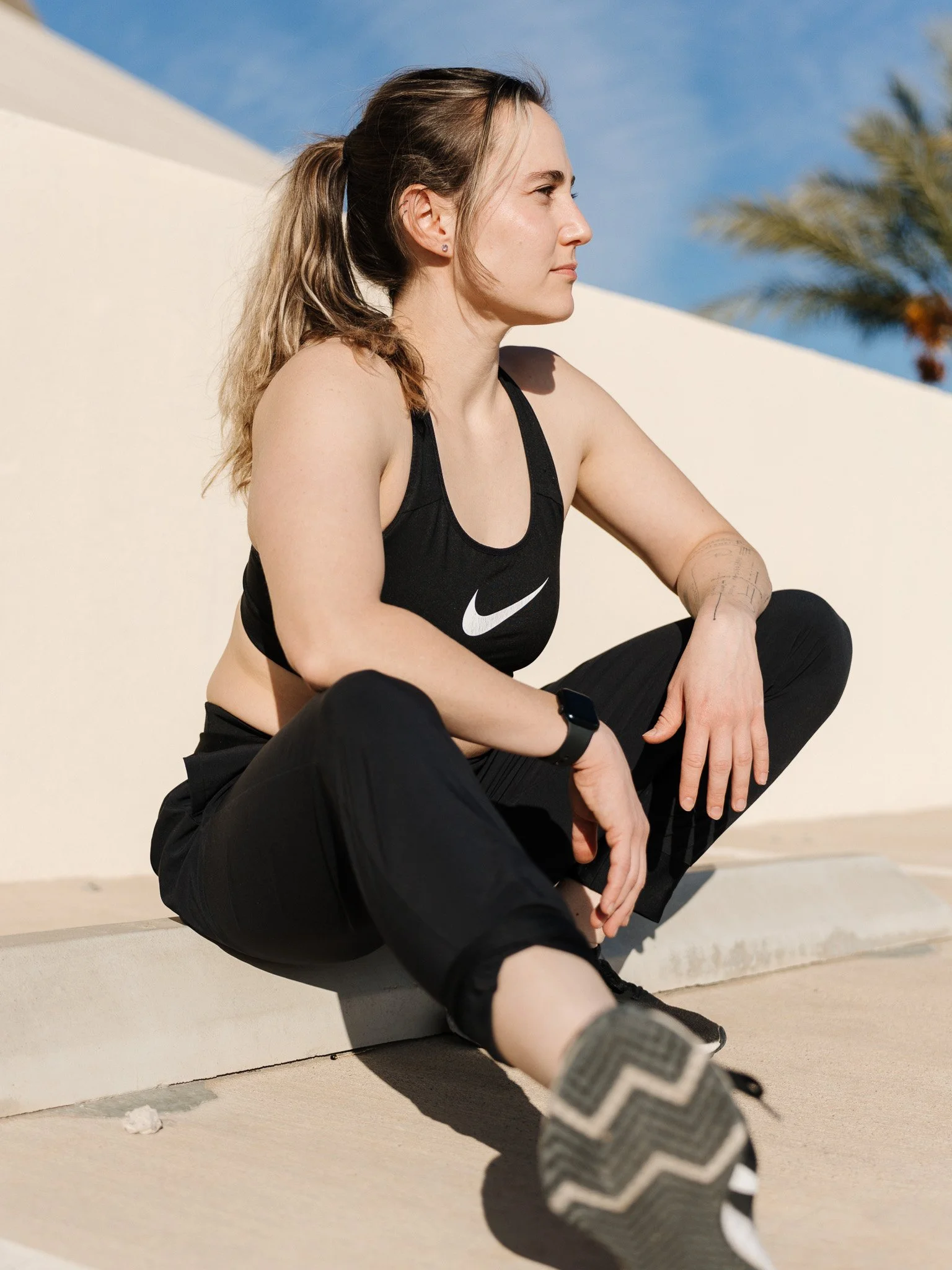 Woman sitting on sidewalk in workout attire, looking to the side, with a palm tree and blue sky in background.