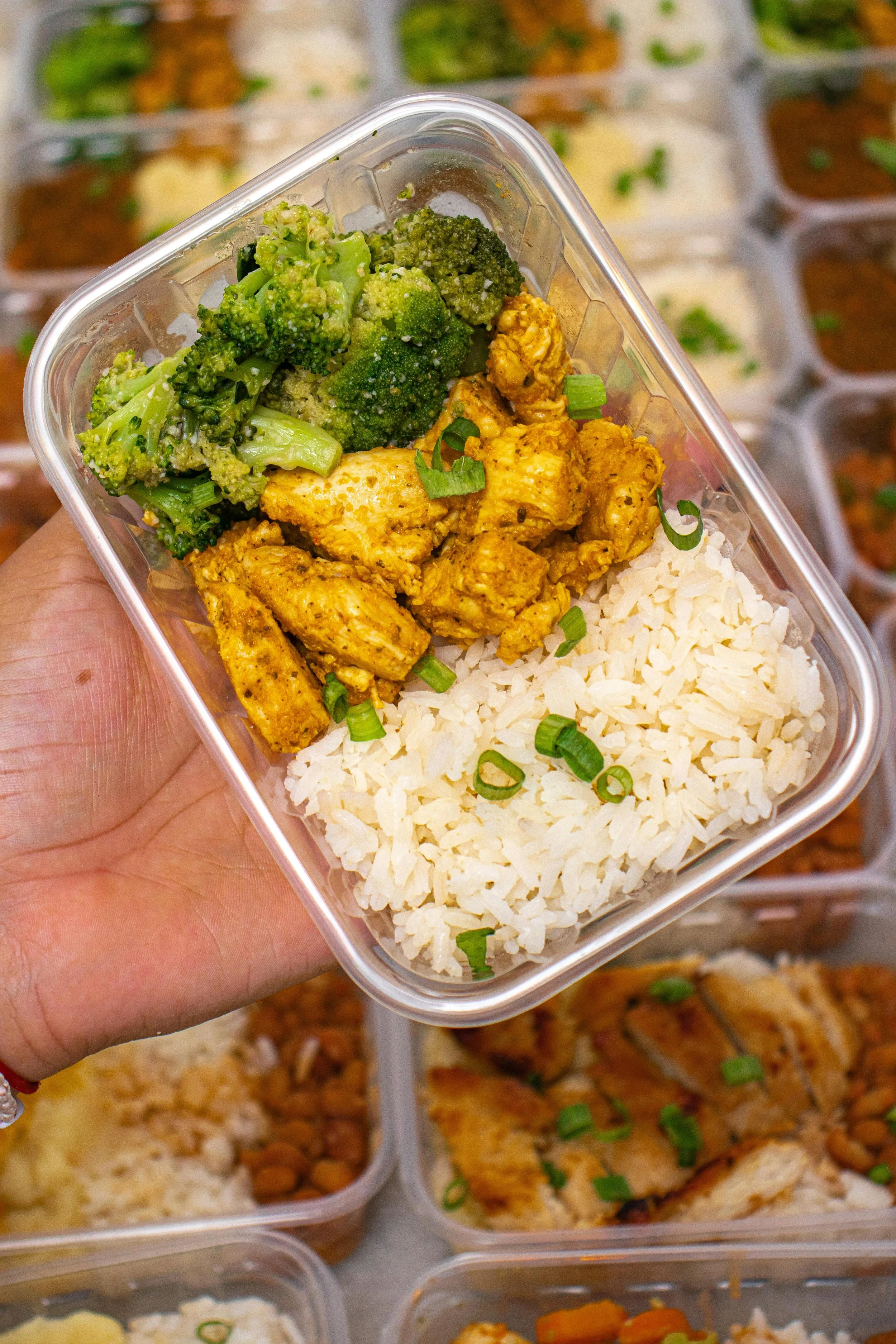 A hand holding a clear plastic container with cooked broccoli, seasoned chicken pieces, and white rice topped with chopped green onions.