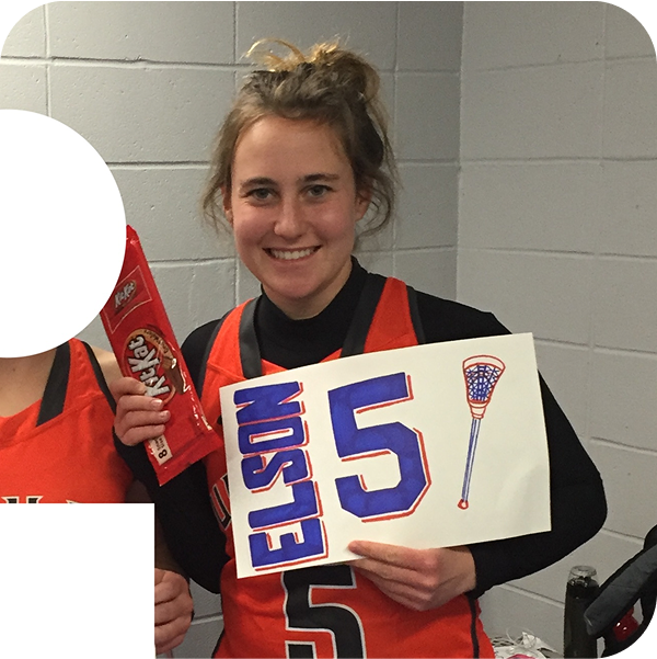 A young woman with blonde hair in a messy bun, wearing a black shirt and red-orange sports jersey, holding a handmade sign that says "ELIST 5" with a blue and red design, and a lacrosse stick drawing. She is smiling and standing against a gray cinder block wall.