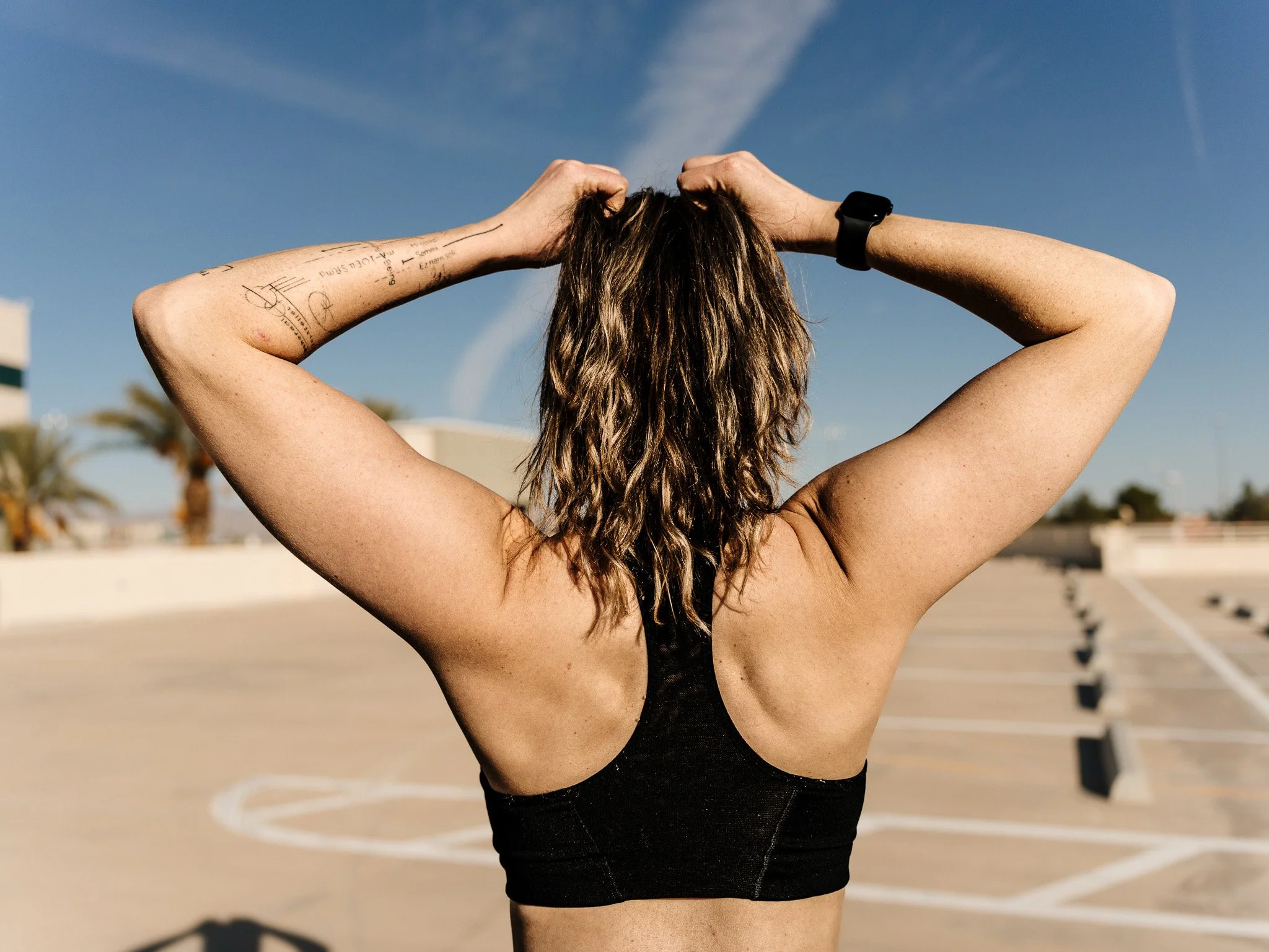 Back view of a woman with wavy brown hair wearing a black sports bra, standing in an empty parking lot with palm trees and a clear blue sky.