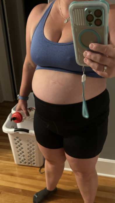 A woman taking a mirror selfie in workout clothes, holding a water bottle, with a laundry basket and hardwood floor in the background.