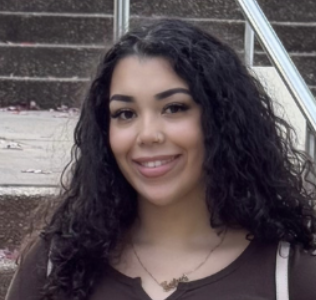 A young woman with curly dark hair smiling outdoors, standing near stone steps and a metal handrail.