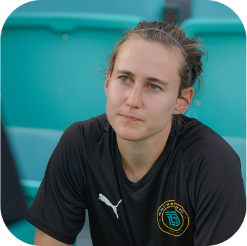 A young woman with brown hair tied back, wearing a black Puma sports shirt with a logo, sitting outdoors near a turquoise background.