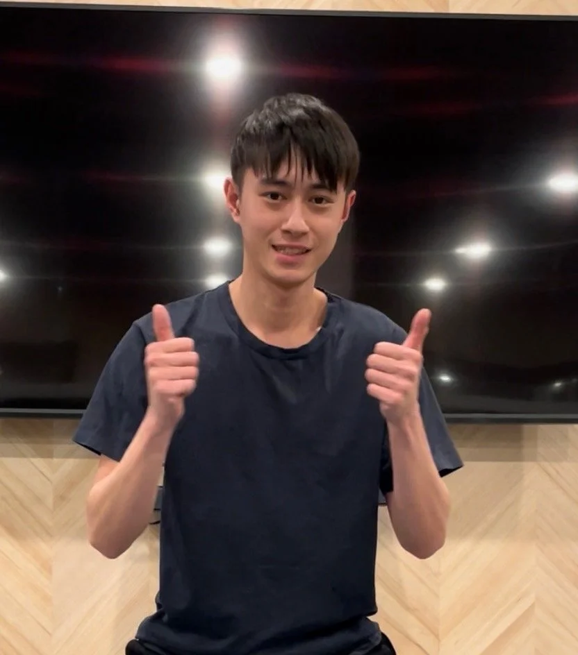 Young man giving two thumbs up, smiling, in front of a large flat-screen TV on a wall