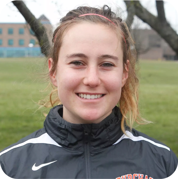 A young woman with blonde hair wearing a black Nike athletic jacket with white stripes on the shoulders, smiling outdoors with trees and a grassy field in the background.
