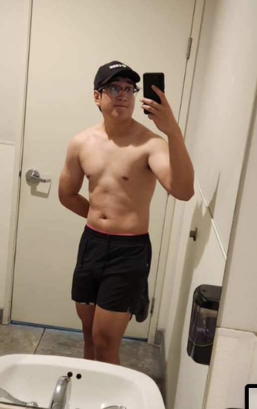 A shirtless young man taking a mirror selfie with a smartphone in a bathroom. He is wearing a black cap, glasses, and black shorts.