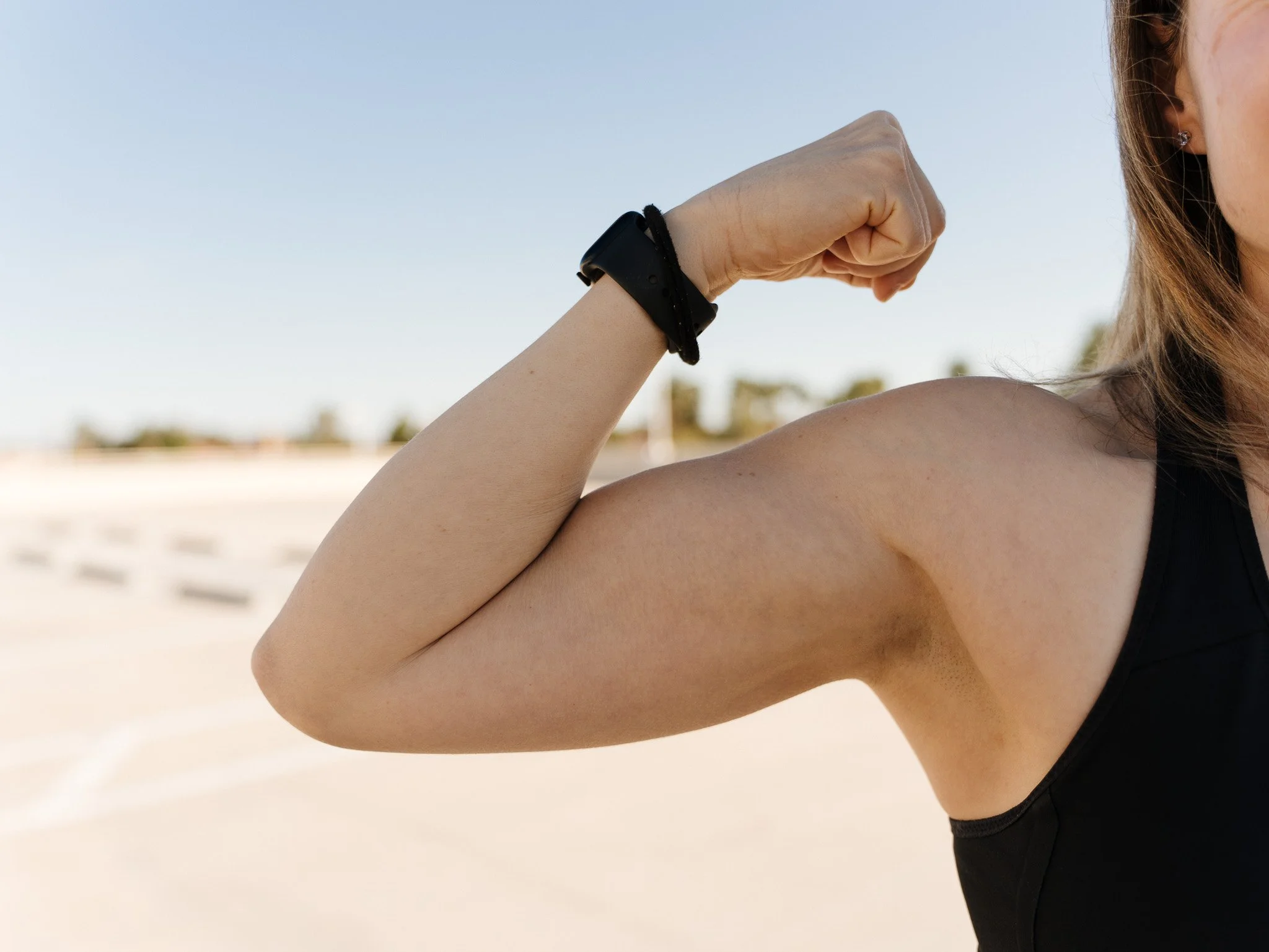 Close-up of a woman flexing her right arm to show her bicep muscle, outdoors with a blurred background of a sunny landscape and sky.