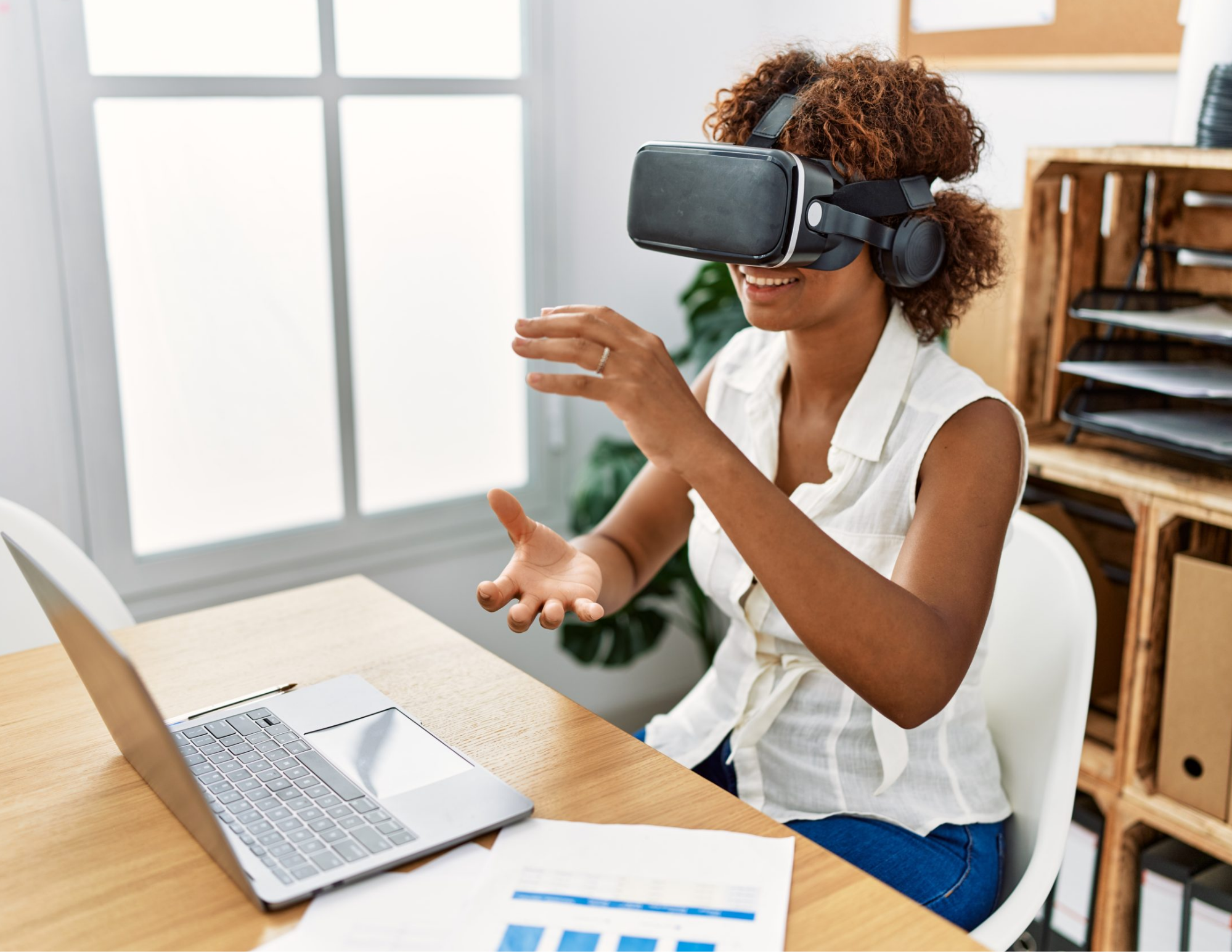 Woman wearing a virtual reality headset and gesturing beside a laptop at a desk