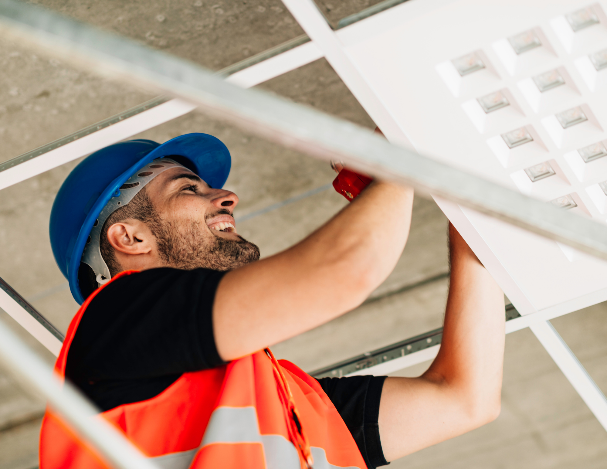 A man wearing a blue safety helmet and an orange safety vest is working underneath a white ladder or scaffolding. The photo is taken from below, with the man smiling and reaching upwards.