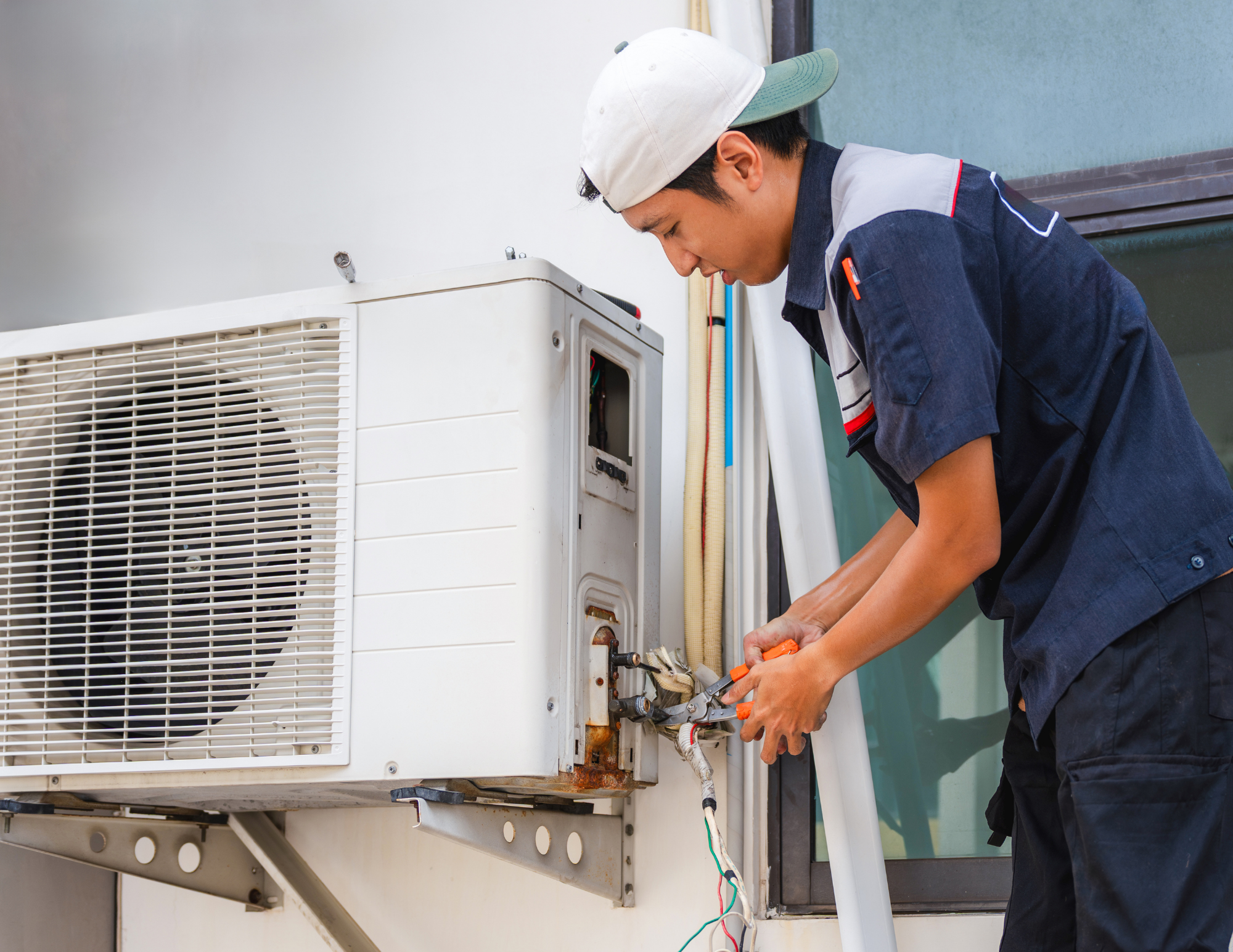 A technician wearing a white cap and dark uniform repairing an air conditioning unit outside a building.