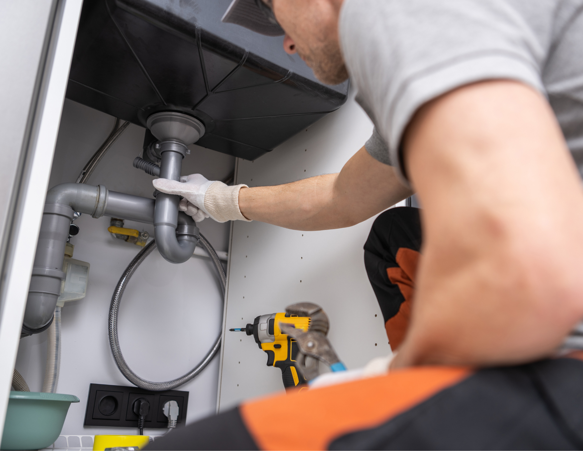 A person working underneath a sink, wearing gloves, using a cordless drill, with plumbing pipes visible and a wall socket nearby.