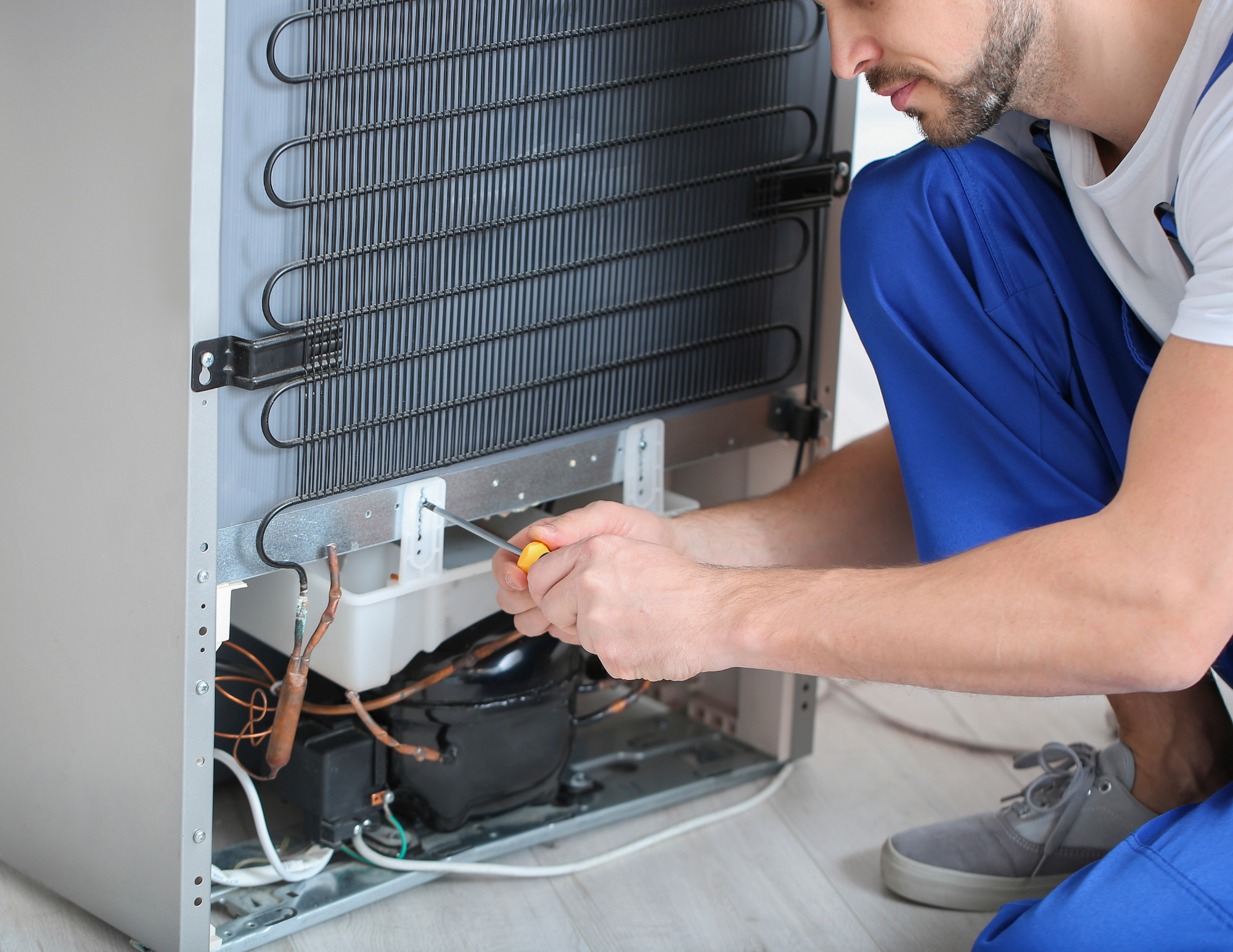 A technician fixing or maintaining an air conditioning unit or refrigerator compressor inside a metal cabinet, using a screwdriver.
