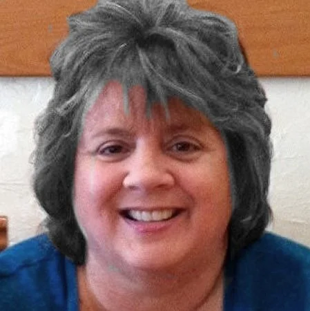 A smiling middle-aged woman with gray hair, wearing a blue top, sitting indoors.