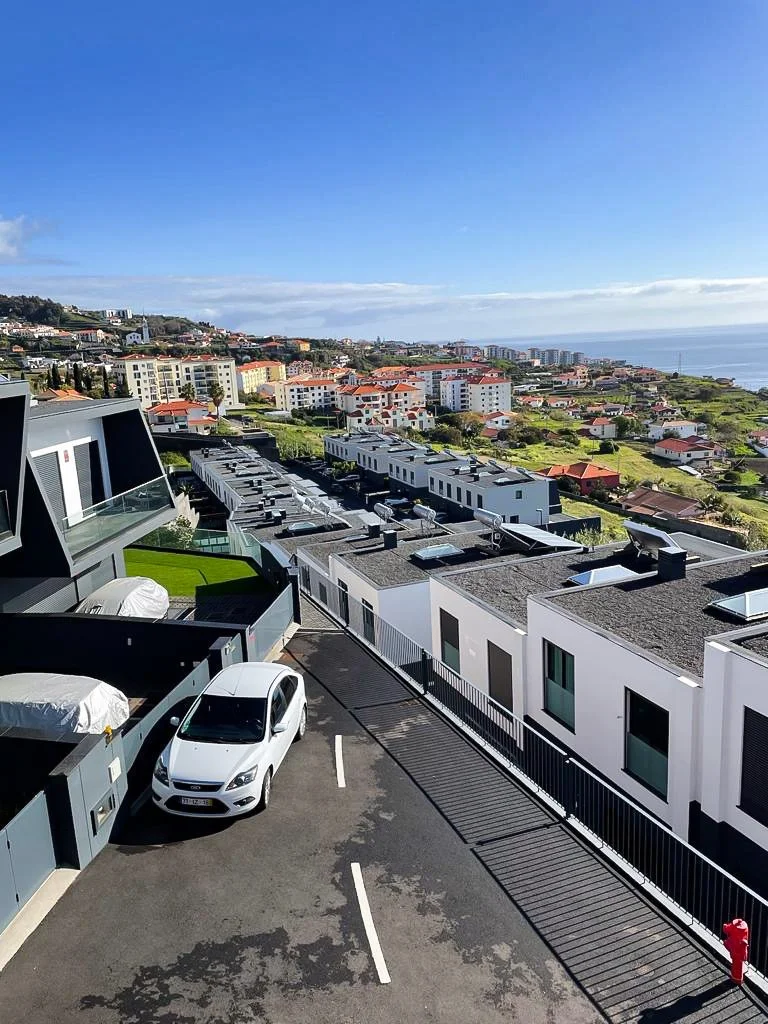Modern residential buildings overlooking a coastal city with a parking lot, white car, and scenic ocean view under a blue sky with some clouds.