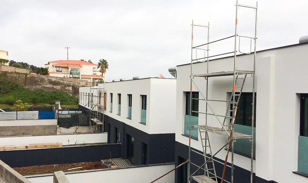 Construction site with scaffolding around a modern white and gray building, ongoing building work, and residential houses in the background.