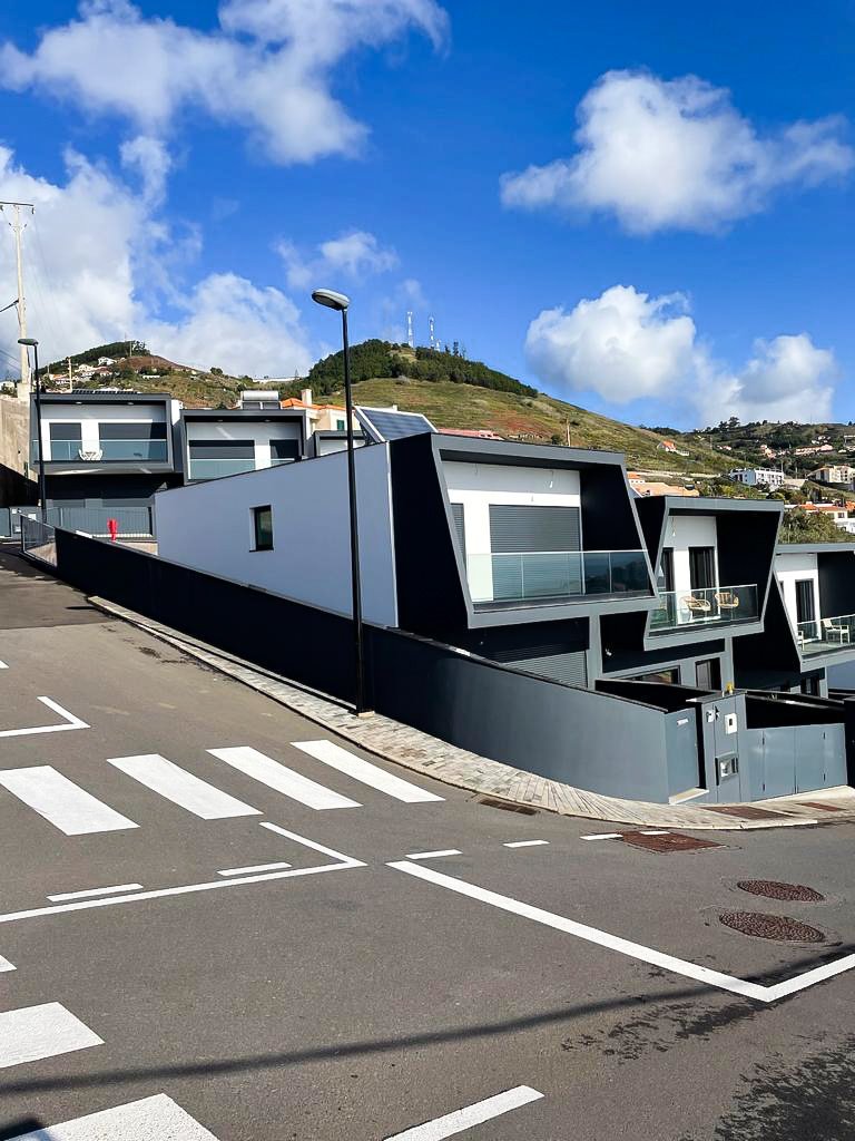 Modern white and black residential buildings with glass balconies on a sloped street, under a partly cloudy blue sky, with hillside in the background.