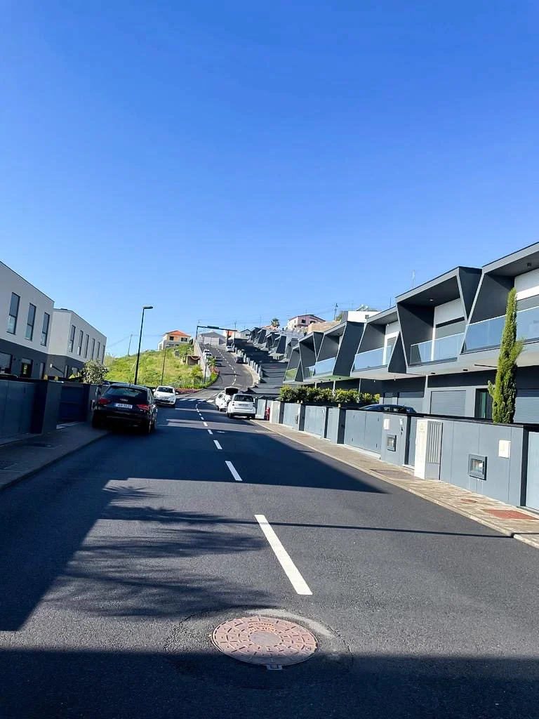 Residential street with modern houses on the right and parked cars along the curb, under a clear blue sky.