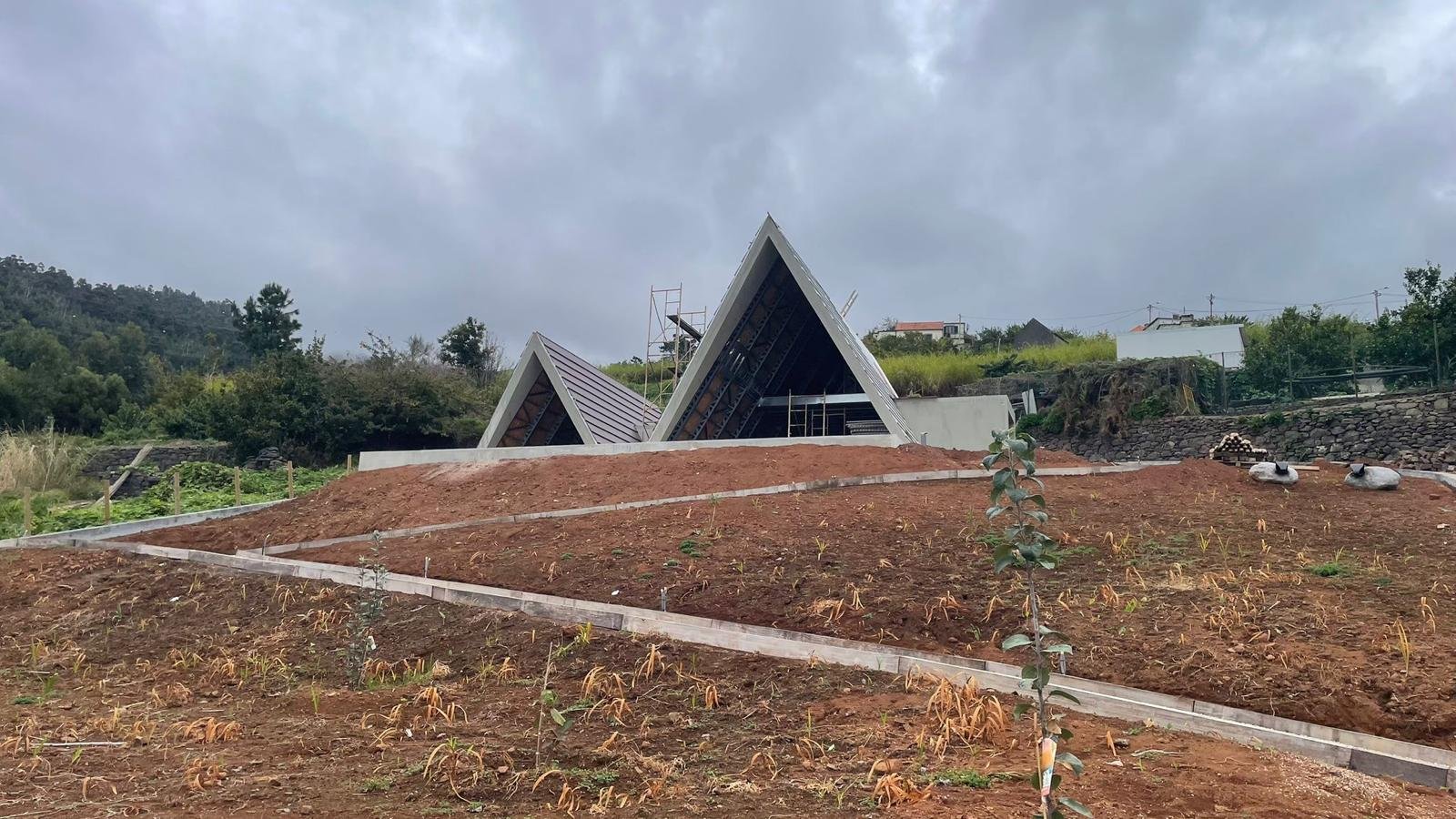 Construction site with modern angular building structures in the background, and a sloped, tilled, reddish-brown soil lot with small plants in the foreground.