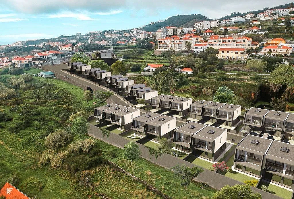 A hillside housing development with multiple modern, box-shaped houses in a neat row, overlooking a lush, green landscape with trees and smaller houses in the distance.