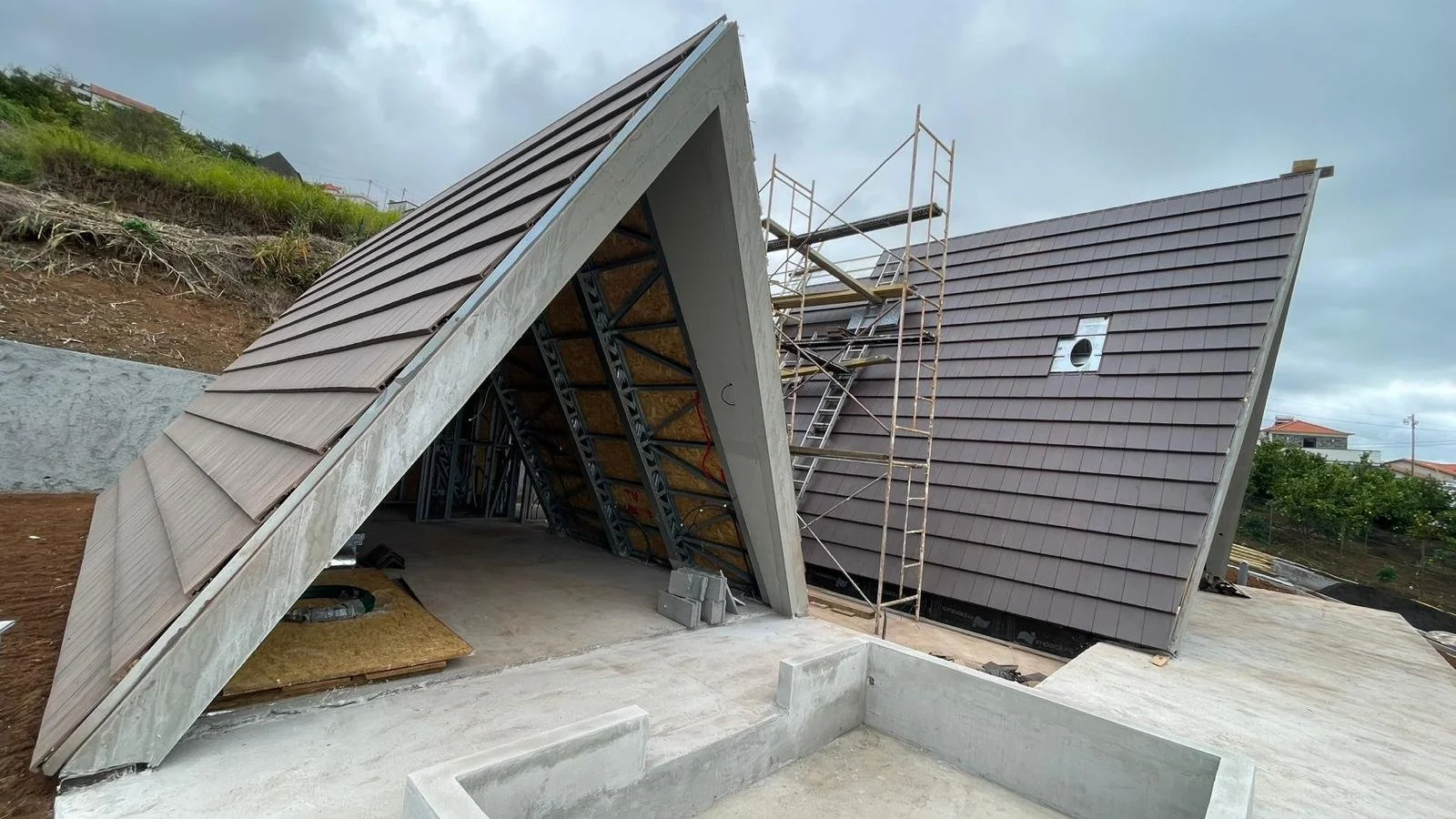 Under-construction modern house with angular roofs, scaffolding, and exterior wall panels, with a cloudy sky and nearby houses in the background.