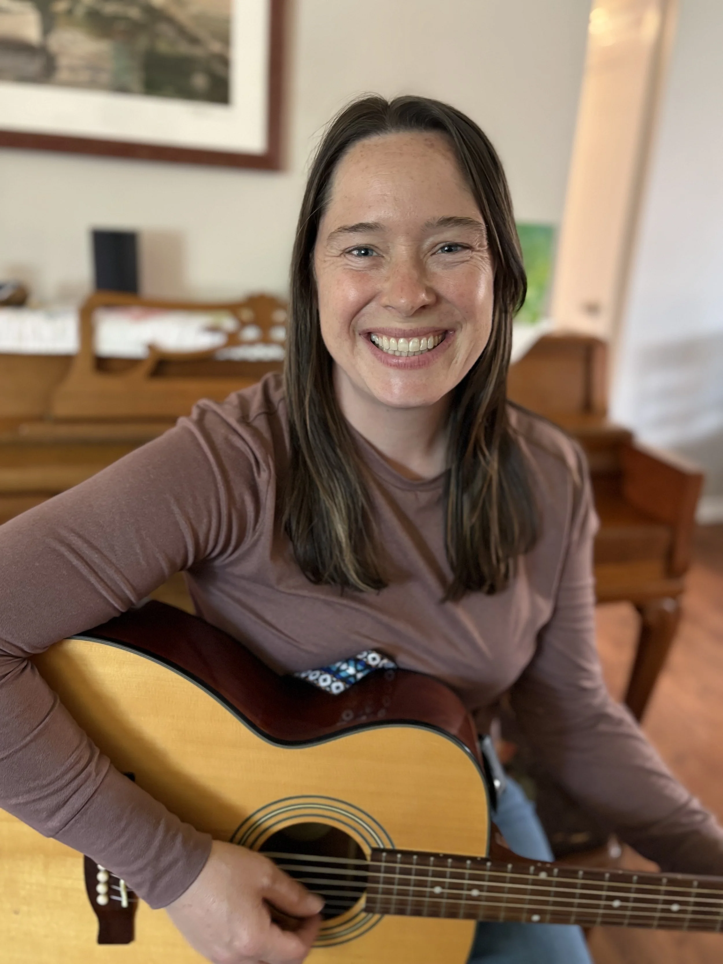 A woman with long brown hair playing an acoustic guitar, sitting in a room with a framed picture on the wall behind her.