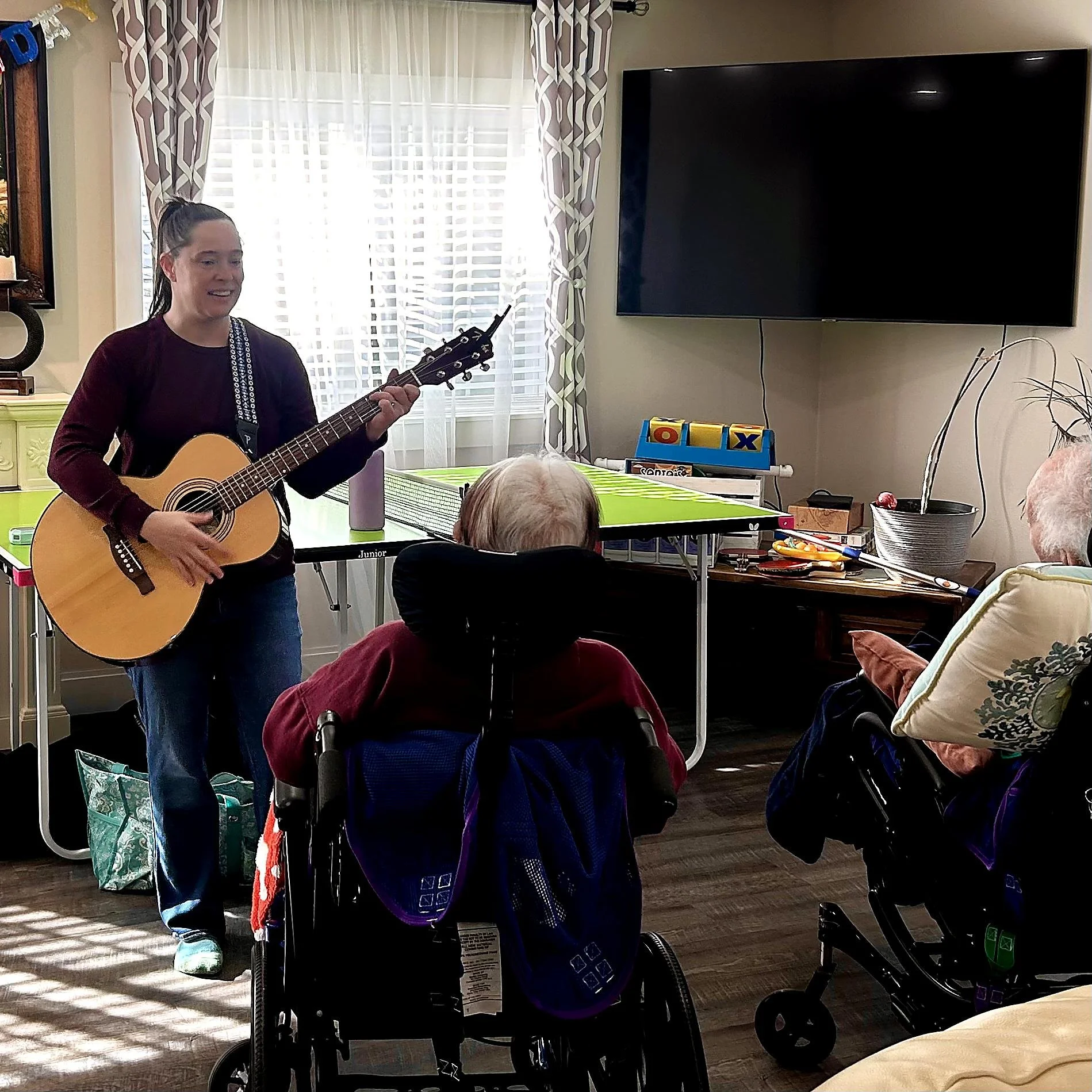 A woman playing an acoustic guitar in a brightly lit room with striped curtains, multiple wheelchairs and elderly people watching.