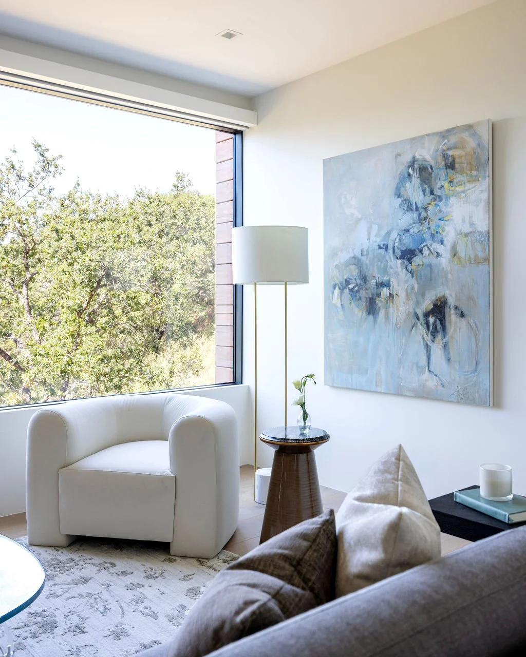 Bright living room with large window showing trees outside. Includes a white armchair, a tall floor lamp, a small round side table with a flower, a beige sofa with pillows, and abstract artwork on the wall.